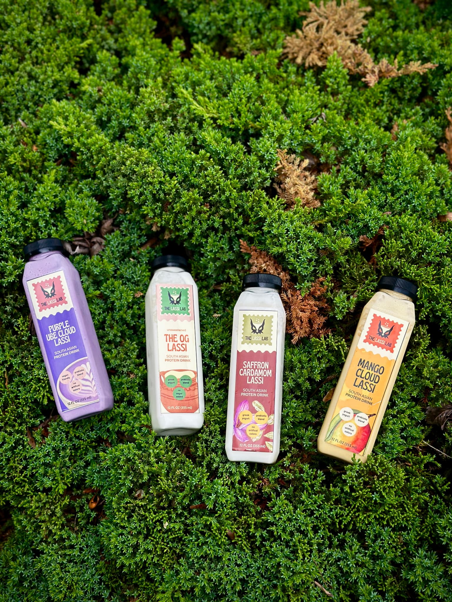 The four Lassi Lab bottles — purple ube, the OG, saffron cardamom, and mango cloud — laid out on moss
